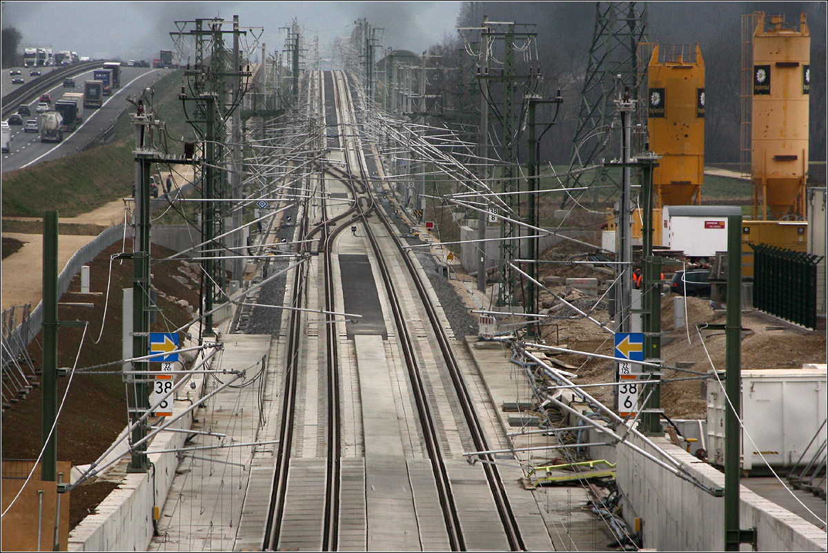 Der elegante Schwung der Gleise und die Dichte der Oberleitungsanlagen - 

Die Gleise passen sich dem hügeligen Geländeprofil an, was sich an den zahlreichen Neigungswechsel zeigt. Die ersten Schnellfahrstrecken durchstießen die Landschaft in fast gerade Linie, die neueren Strecken hingegen zeigen ein Auf und Ab, wodurch weniger Erdbewegungen notwendig sind. Die TGV-Strecken in Frankreich sind ähnlich trassiert, mit relativ wenigen Tunnels.

Nur ganz schemenhaft ist der Maschendrahtzaun erkennbar, durch den ich hier fotografiere. Ich stehe dabei auf dem östlichen Tunnelportal des ca 8,2 km langen Albvorlandtunnels. Bis vor kurzem war diese Stelle noch Baustellenbereich, doch inzwischen ist die Straße wieder öffentlich zugänglich und der große Baustellenbereich wird wieder renaturiert. 

16.02.2022 (M)


