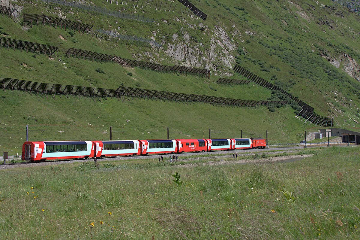 Der Glacier-Express wird als langsamster Schnellzug der Welt bezeichnet. Heutzutage wird er mit modernen Panoramawagen f�r touristische Zwecke betrieben.
Habe hier einen Nachschuss auf die Komposition am 05.07.2026 kurz vor dem Oberalppass aufgenommen. Am Hang sind Lawinenverbauungen zu erkennen. Bald f�hrt der Zug in die Lawinengalerie ein. Diese Bauten machten es m�glich, auch im Winter von Andermatt nach Disentis zu fahren.