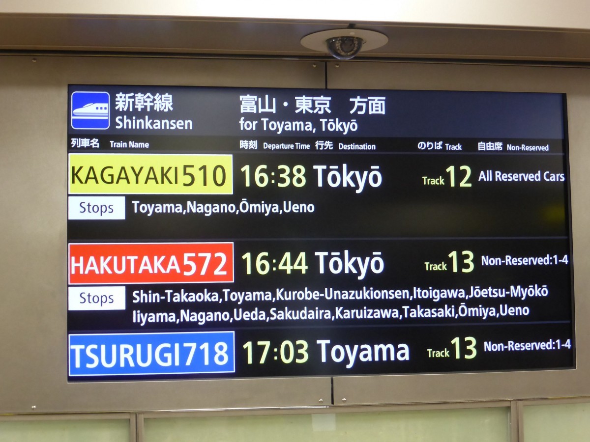 Der Hokuriku-Shinkansen: Die übersichtliche Anzeigetafel der Abfahrten im Bahnhof Kanazawa. Mattbildschirm. Die Tafel leuchtet abwechselnd auf Englisch und Japanisch. Die drei Zugsgattungen in unterschiedlicher Farbe. Als Detail fällt die Unterscheidung zwischen langem und kurzem O auf (langes O mit Längsstrich). Die Angabe der Position der Türen ist hier nicht nötig, weil diese auf Tafeln direkt am Gleis angebracht sind. 27.März 2015.   
