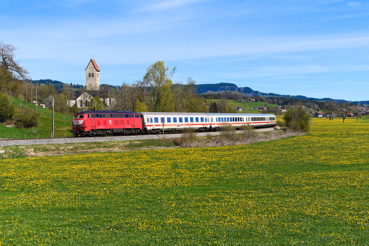 Der IC 2084 wurde gegen Ende seiner Einsatzzeit tatsächlich zumeist im Wendezug-Betrieb gefahren. Zwischen Immenstadt und Augsburg fielen daher die gewohnten Motiv-Klassiker für Aufnahmen des Zuges weg, allerdings boten sich neue Varianten, sofern man wie ich mit einem  Nachschuss  gut leben kann. Am 19. April 2025 war die orientrote 218 406 für den  Nebelhorn  eingeteilt. Bei Stein im Allgäu konnte ich sie mit ihrem kurzem Fernzug bei fast voller Löwenzahnblüte fotografieren. Nur die Bewölkung am Himmel hätte etwas schöner sein dürfen.   