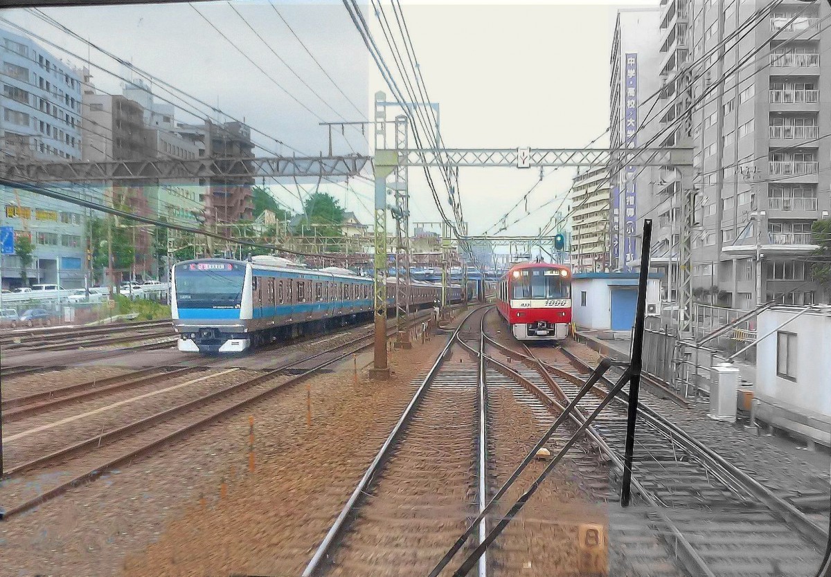 Der Keihin Kyûkô-Konzern (auch Keikyû-Konzern genannt), Grundangaben: Dieser grosse Konzern mit über 9000 Angestellten verbindet Tokyo mit Yokohama. Anders als zwischen Tokyo und Yokohama die JR mit nur wenigen grossen Stationen und Fern-sowie Güterzügen auf ihren Gleisen führt der Keikyû-Konzern eng der Küste entlang durch ein Gewirr von bescheidenen Siedlungen, Kleingewerbe und Fabrikanlagen. Zwischen Tokyo und Yokohama wird die Industriestadt Kawasaki bedient. Von Yokohama aus fahren die Keikyû-Züge dann auf die Miura-Halbinsel hinaus, welche das Westufer der Bucht von Tokyo bildet. Alle Strecken des Keikyû-Konzerns sind normalspurig (1435 mm), 1500 V Gleichstrom. Ausser der Hauptlinie gibt es zahlreiche Zweiglinien, insgesamt 87 km Strecke. Keikyû-Züge fahren zudem über die Städtische U-Bahn Tokyo und über wichtige Normalspurstrecken nordöstlich von Tokyo bis zum Internationalen Flughafen Narita. Der Name Keihin Kyûkô bedeutet  Schnellverbindung zwischen Tokyo und Yokohama . Die Strecke war bereits 1899 elektrisch. Bild: Ein normalspuriger roter Keikyû-Zug (Zug 1033) neben einem 1067mm-Spur-Zug der JR (Serie 233) zwischen Tokyo und Yokohama, vor Kanagawa, 25.Juni 2011. (Aufnahme durch die verglaste Führerstandrückwand eines Gegenzugs)