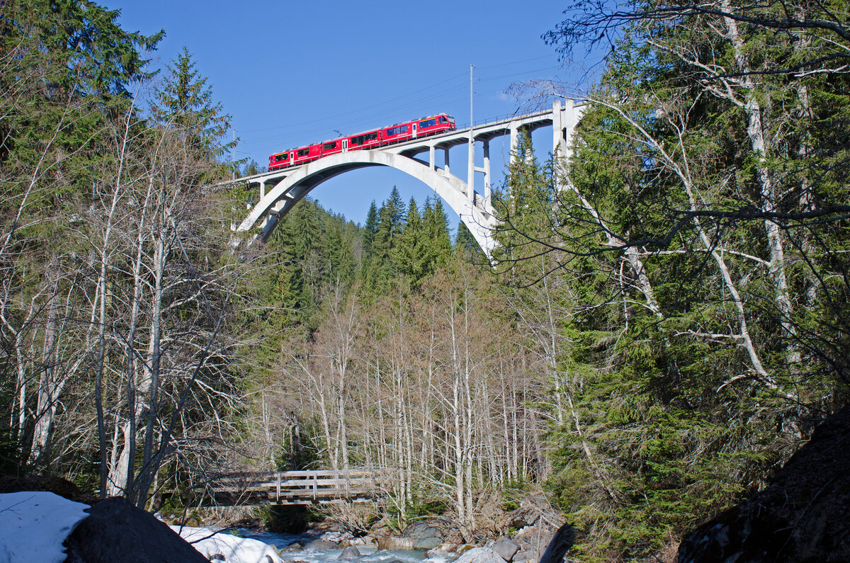 Der Langwieser-Viadukt bei Langwies (Bauzeit. 1.8.1912 bis 1.7.1914) auf der RhB Strecke Chur – Arosa hier von der Bergseite gesehen. Bogenspannweite 100 m, Laenge 287 m, Hoehe 62 m. 1914 war es die bedeutendste Bahn-Eisenbeton-Bogenbruecke der Welt. Hier befaehrt der Regio 1425 (Chur ab 09:08 Uhr, Arosa an 10:09 Uhr) mit dem Triebzug der Serie 35.. um 10:05 Uhr die Bruecke bei der Bergfahrt. Dieser Zug hat eine Verspaetung von zirka 15 Minuten. Diese Aufnahme entstand am 19.04.2022 am Bachbett des Sapuener Bach der unterhalb der Bruecke in die Plessur fliest. Die ABe 8/12 sind dreiteilige elektrische Triebzuege welche die Rhaetische Bahn (RhB) bei Stadler Rail bestellt hat.