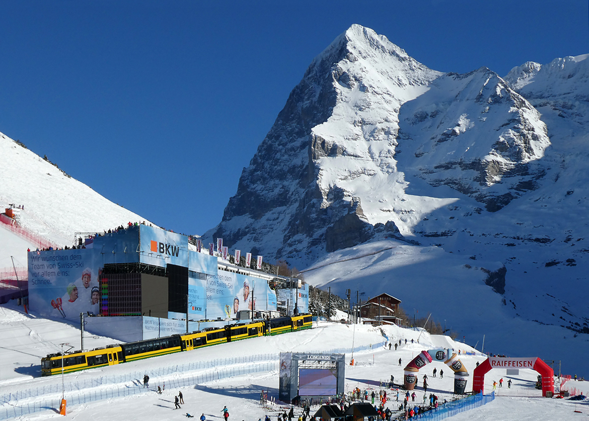 Der Lokalzug von Lauterbrunnen - Wengen nach Kleine Scheidegg fährt in die Station Wengernalp mit dem braunen Stationsgebäude ein. Links ist die Sponsorentribüne der Lauberhornabfahrt zu sehen. Auf dem grossen Bildschirm unten in der Mitte können die Zuschauer das Skirennen verfolgen. Die beiden grossen Werbebogen rechts bringen zusätzlich Farbe ins Spiel. Im Hintergrund ist der Eiger mit der weltberühmten Eigernordwand zu sehen. Wengernalp, 11.1.2024