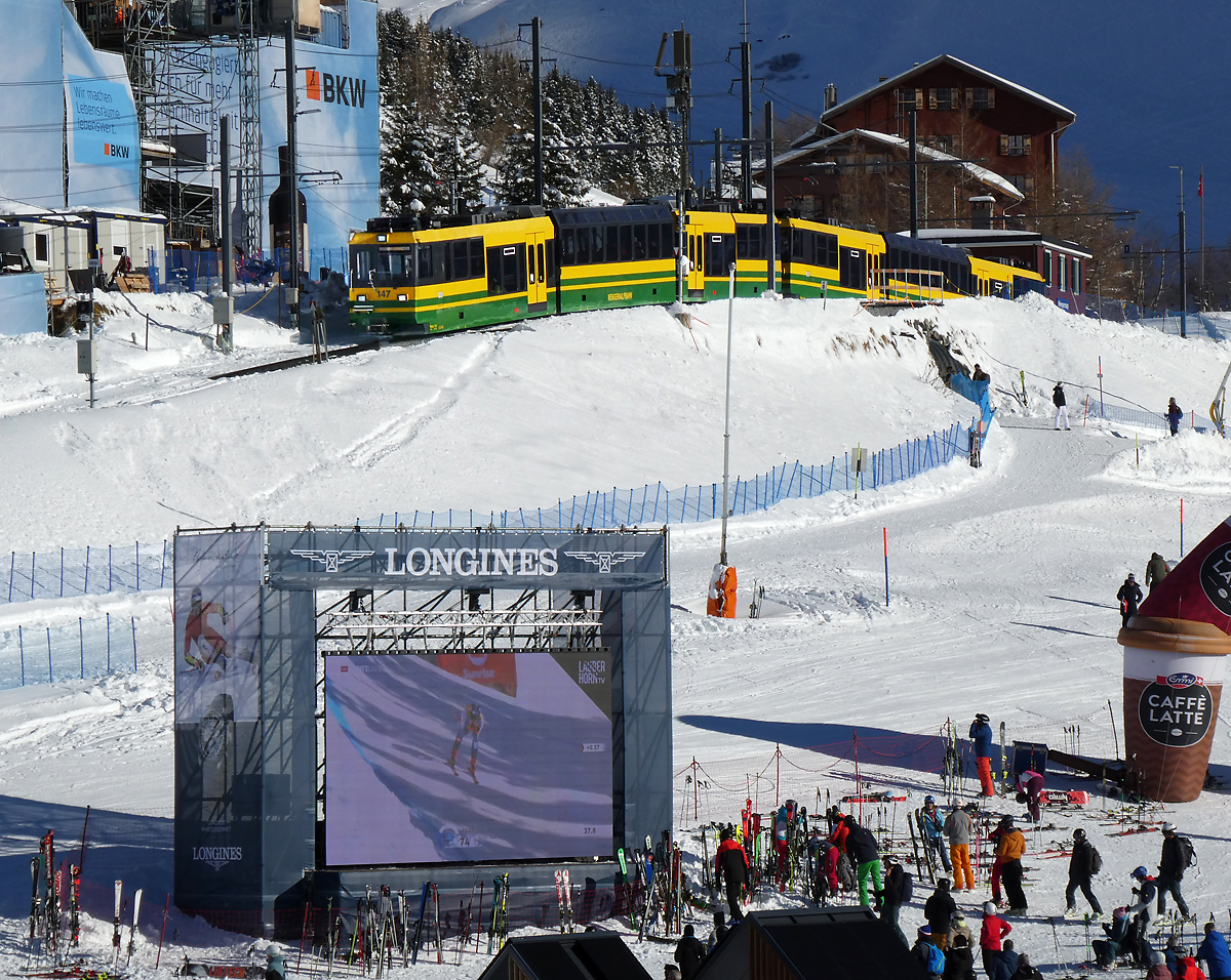 Der Lokalzug von Lauterbrunnen - Wengen fährt in die Station Wengernalp mit dem braunen Stationsgebäude ein. Auf dem grossen Bildschirm können die Zuschauer das Skirennen verfolgen. Wengernalp, 11.1.2024
