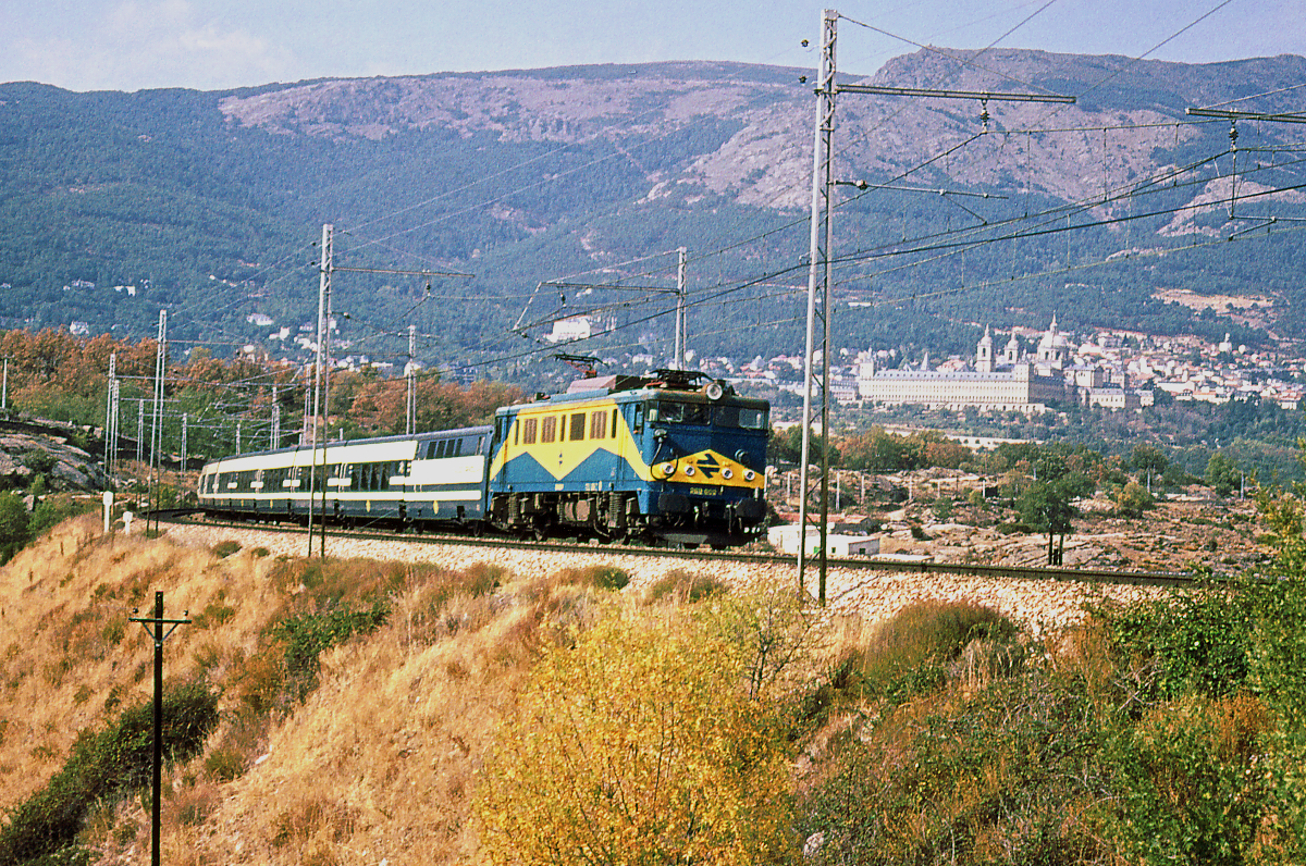 Der nachmittägliche Talgo Madrid - Gijon ist auf dem Weg durch die Sierra de Guadarrama, dem Gebirge, welches die spanische Hauptstadt von den weiten Ebenen Altkastilliens trennt. Im Hintergrund liegt das nkönigliche Kloster El Escorial. Der Talgo 30131 hat Madrid Chamartin um 14.40 verlassen und wird Gijon um 21.59 erreichen. Es führt die Lokomotive 269 602, mit Chopper-Steuerung, gebaut von Mitsubishi in Japan. Oktober 1983