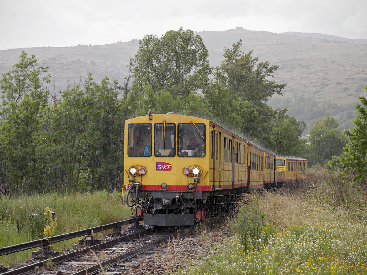 Der  Petit Train Jaune  (klener gelber Zug) der  Linge de Cerdange  fährt am ehemaligen Bahnhof Ur Les Escaldes am 02.07.2019 vorbei. Der plötzlicher Sommerregen verwirrte meine Fotopläne für diesen Zug. Ich konnte nur dieses eine Foto machen.