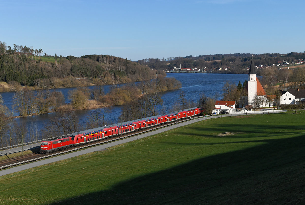 Der RE 4074 von München HBF nach Passau wurde am 15. März 2020 als  Sandwich -bespannte Doppelstockwagengarnitur geführt. Bei Hausbach hat der Zug sein Ziel bald erreicht. In flotter Fahrt geht es am Ufer der Donau entlang und die älteste niederbayerische Rundkirche wird passiert. Am Zugschluss befand sich die gut im Lack stehende 111 022, inzwischen eines der ältesten elektrischen Triebfahrzeuge, welche die Deutsche Bahn im Bestand hat.  