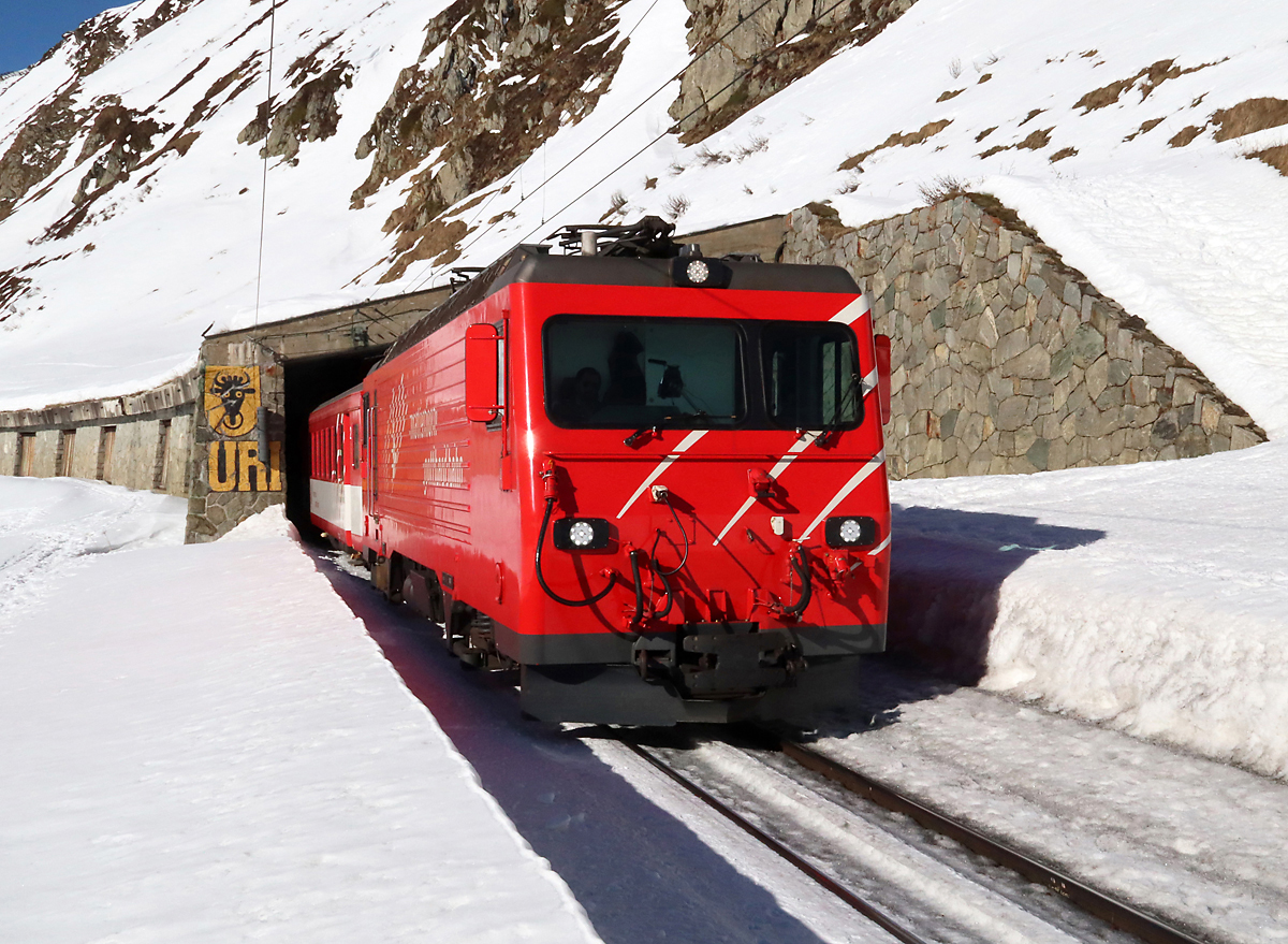 Der Regionalzug von Andermatt nach Disentis verlässt den Galerietunnel kurz vor der Station Oberalppass (2033 müM). Hier in der Nähe befindet sich die Kantonsgrenze Graubünden/Uri, siehe das Kantonswappen von Uri mit dem Uri-Stier am Tunnelportal. Oberalppass, 13.2.2023