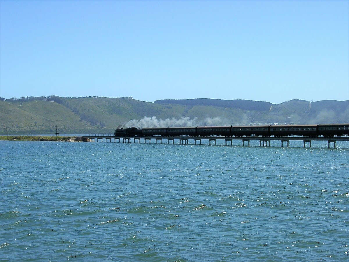 Der täglich verkehrende Outeniqua Choo Tjoe von George nach Knysna als letzter regelmäßige Dampfzug Südafrikas auf der Knysna Lagoon Brücke. 2006 zerstörten heftige Regenfälle, Erdrutsche und Überschwemmungen wichtige Teilstrecken zwischen George und Knysna, deren Instandsetzung zu teuer waren und dieses Teilstück daher früh außer Betrieb genommen wurde.
Aufnahme am 15.11.2002 von einer Schiffsrundfahrt auf der der Knysna Lagoon.