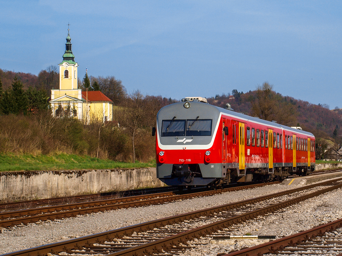 Der Triebwagen 713 118 fährt am 10.04.2015. als nachmittäglicher RegioExpress 609 von Ljubljana nach Novo Mesto in den Bahnhof von Visnja Gora ein, wo bereits ein Personenzug in der Gegenrichtung auf die Kreuzung mit dem RG wartet. Der VT präsentiert sich im Neulack, da er vor knappen zwei Monaten in der Werkstätte von Ptuj eine Hauptausbesserung erhielt. Links zu sehen ist die Kirche der Hl. Ana.