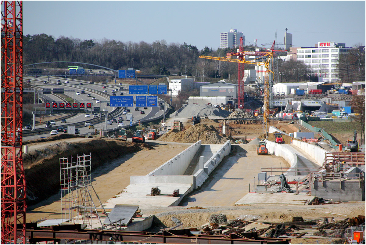 Der zukünftige Abzweig zum Flughafen -

Blick auf die im Rohbau fertigen Tunnelrampen für den Abzweig von der Neubaustrecke Stuttgart - Ulm zum Flughafen. Weiter im Osten wird die Flughafenschleife wieder in die Neubaustrecke eingefädelt.
Weiter hinten ist das Portal zum Fildertunnel zu erkennen, wo die Strecke vom Hauptbahnhof her kommend wieder ans Tageslicht kommt. Zwischen der Autobahn und der linken Rampe wird das Gleis in Richtung Ulm verlaufen, zwischen den beiden Rampen diese in Richtung Stuttgart. 
Auf der anderen Seite der Straßenbrücke auf der ich hier stehe, sollen links und rechts der beiden Hauptgleise zwei weitere Tunnelrampen zu einer weiteren Tunnelstrecke zum S-Bahnhof Flughafen/Messe entstehen über die dann Gäubahn angeschlossen werden soll. Allerdings gibt es da auch inzwischen Überlegungen diese mit einem zusätzlichen 11 km langen Tunnel an den Flughafen-Fernbahnhof anzuschließen. Auf dieser Trasse würden die Züge der Gäubahn fast im Kreis fahren und die Neubaustrecke zwischen Fildertunnel und dem hier sichtbaren Abzweig unterirdisch kreuzen. 
Solche Eigenkreuzungen gibt es bei Schraubentunnels auf Gebirgstrecken. Im ebene Gelände wäre ein Beispiel die Kramerkurve südlich von Berlin, oder die Streckenführung in Tønsberg in Norwegen südlich von Oslo. Oder die Schleife in Rendsburg, hier ähnlich der Schraubtunnels um Höhe zu gewinnen.

27.02.2022 (M)