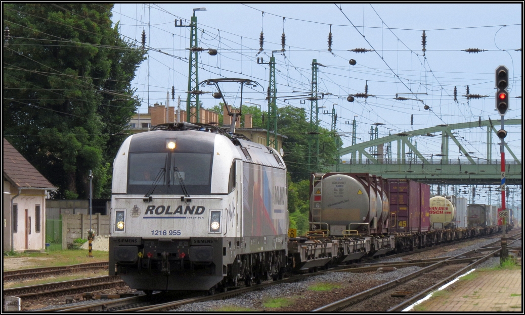 Die 1216 955 der WLC im Roland Spedition Design kommt mit einen Aufliegerzug durch Komárom gefahren. Szenario bildlich festgehalten Anfang August 2015.