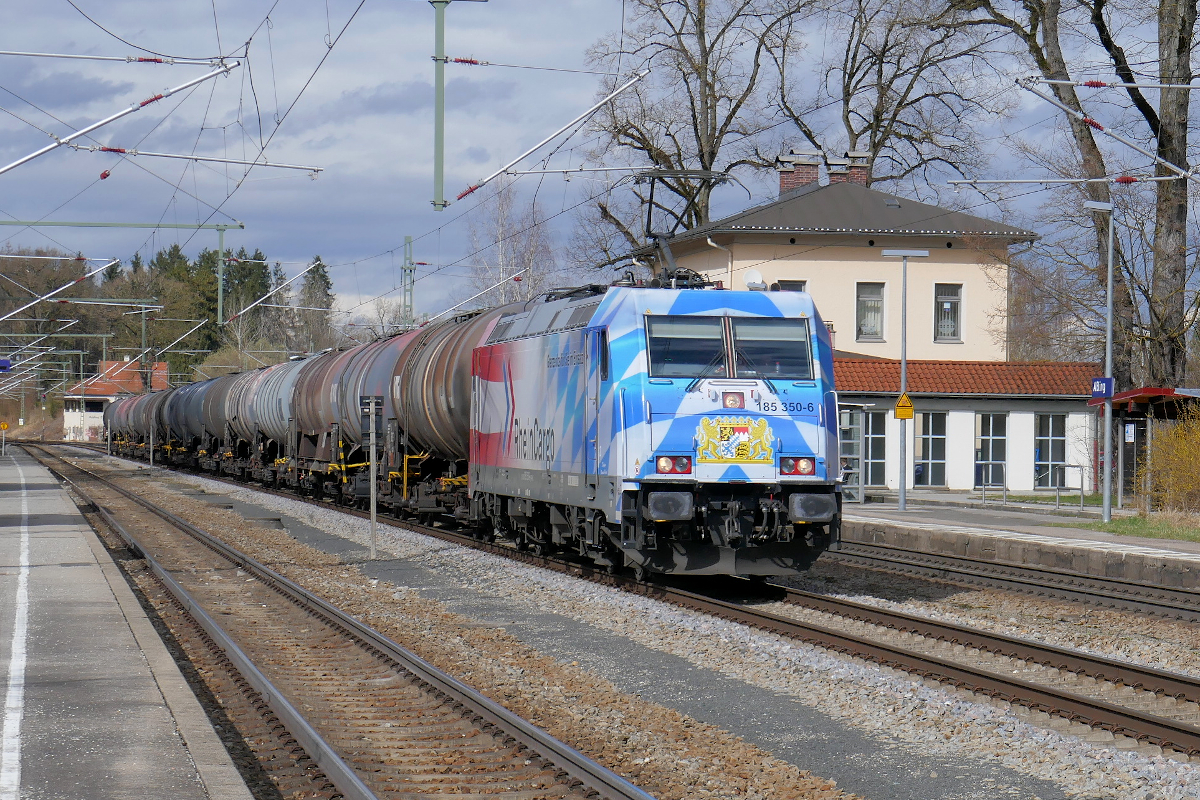 Die 185 350 der Rheincargo ist mit den Bayerischen und Österreichischen Fahnen und Wappen beklebt. Ihre  Bayerische Seite  glänzt in der Sonne des 12. März 2020, als sie mit einem Kesselwagenzug den Bahnhof Aßling in Richtung Rosenheim durchfährt.