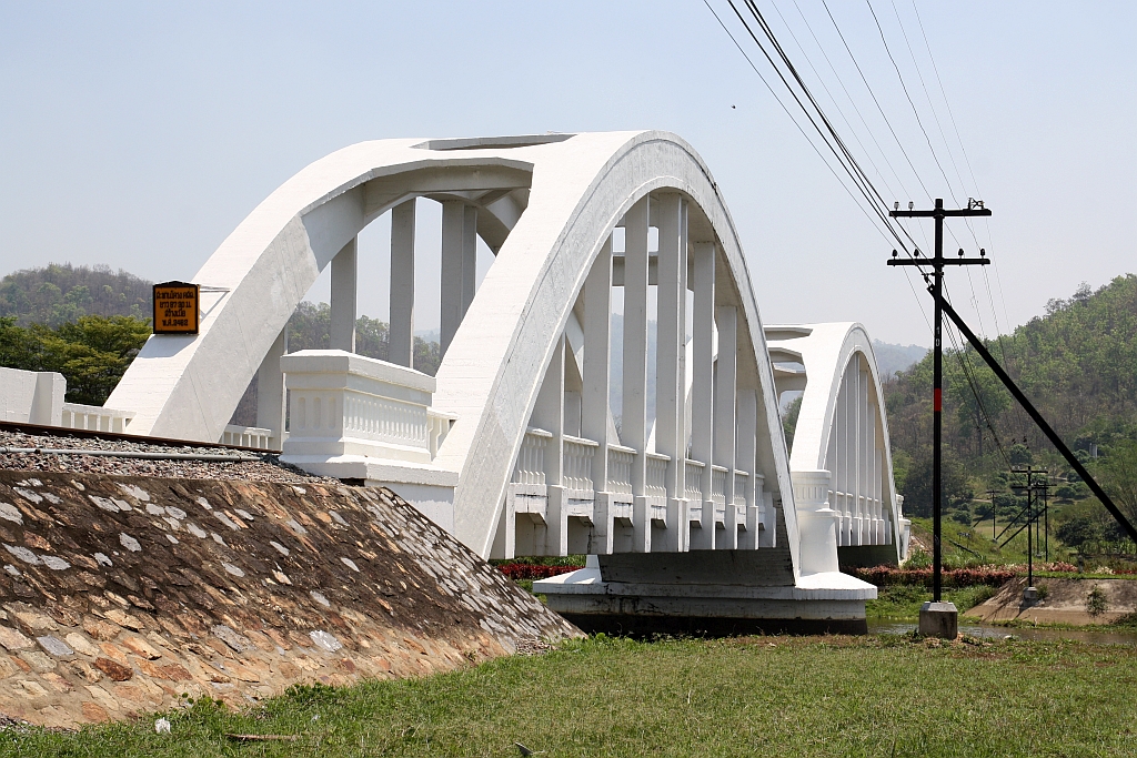 Die 1919 fertiggestellte 87,3m lange Tha Chomphu Bridge über den Mae Nam Tha, von den Einheimischen Saphan Khao (=Weiße Brücke) genannt, gilt als eines der Wahrzeichen der Provinz Lamphun und ist deshalb ein Touristen-Hotspot mit Parkplatz, Informationsstand, Parkanlage und sogar mit einer Treppe auf den Bahndamm. Bild vom 22.März 2023.
