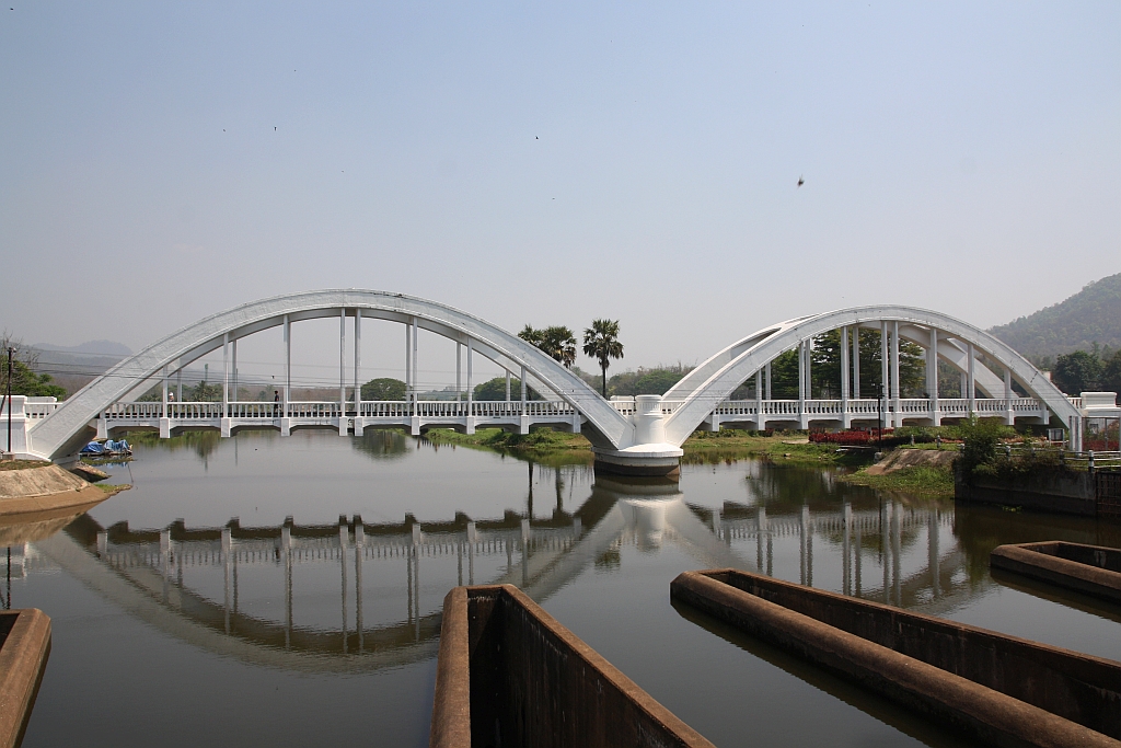 Die 1919 fertiggestellte 87,3m lange Tha Chomphu Bridge über den Mae Nam Tha, von den Einheimischen Saphan Khao (=Weiße Brücke) genannt, gilt als eines der Wahrzeichen der Provinz Lamphun und ist deshalb ein Touristen-Hotspot mit Parkplatz, Informationsstand, Parkanlage und sogar mit einer Treppe auf den Bahndamm. Bild vom 22.März 2023.