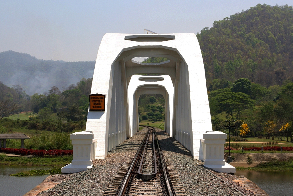 Die 1919 fertiggestellte 87,3m lange Tha Chomphu Bridge über den Mae Nam Tha, von den Einheimischen Saphan Khao (=Weiße Brücke) genannt, gilt als eines der Wahrzeichen der Provinz Lamphun und ist deshalb ein Touristen-Hotspot mit Parkplatz, Informationsstand, Parkanlage und sogar mit einer Treppe auf den Bahndamm. Bild vom 22.März 2023.