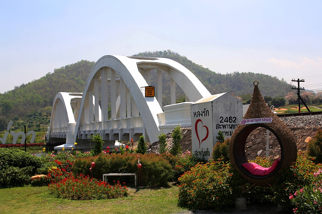 Die 1919 fertiggestellte 87,3m lange Tha Chomphu Bridge über den Mae Nam Tha, von den Einheimischen Saphan Khao (=Weiße Brücke) genannt, gilt als eines der Wahrzeichen der Provinz Lamphun und ist deshalb ein Touristen-Hotspot mit Parkplatz, Informationsstand, Parkanlage und sogar mit einer Treppe auf den Bahndamm. Bild vom 22.März 2023.