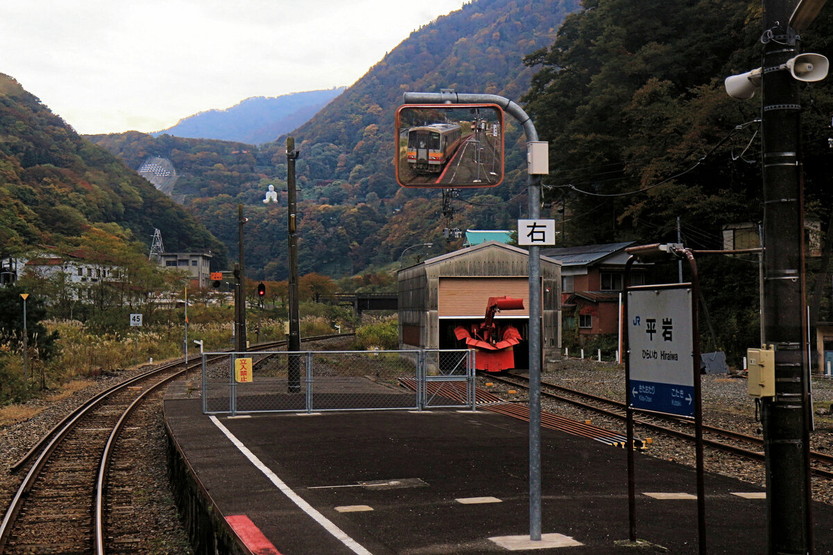 Die Ôito-Linie von JR West Japan: Tief im einsamen Gebirge hält Schienenbus KIHA 120-329 in Hiraiwa, im Spiegel zu sehen. Ein Schneeräumungsgefährt streckt seine Nase aus einem kleinen Schuppen, während auf dem Berg oben ein grosser Buddha die Szene betrachtet. 4.November 2018  
