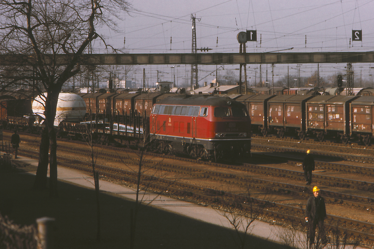 Die 218 223-6 des Bahnbetriebswerkes Mühldorf hat soeben einen Güterzug aus Richtung München-Pasing nach München Laim Rangierbahnhof gebracht. Im Jahr 1975 war die Baureihe 218 eine moderne Lokomotive und der Rangierbahnhof München Laim war noch in Betrieb. In den 1990er Jahren ging der Rangierbahnhof München Nord in Betrieb, Laim wurde in ein Wohnviertel verwandelt, auf den verbliebenen Gleisen werden ICE abgestellt, TXL ändert dort die Bespannung seiner Güterzüge. Seit dem Jahr 2019 wird in Laim an der zweiten Stammstrecke gebaut.
München Laim Rbf, 11. Februar 1975