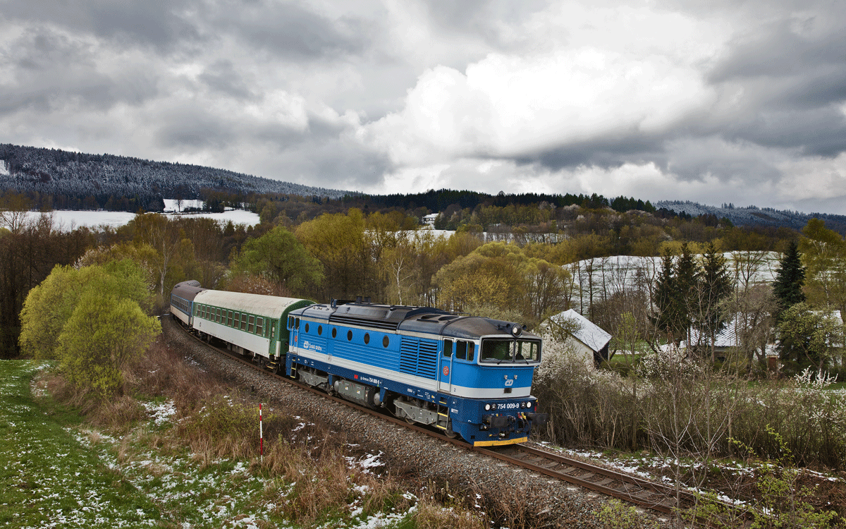 Die 754 009-9 dieselt mit dem Rx 775 unter schneeebeladener Wolkenstimmung in Desenice vorüber.Bild vom 19.4.2017