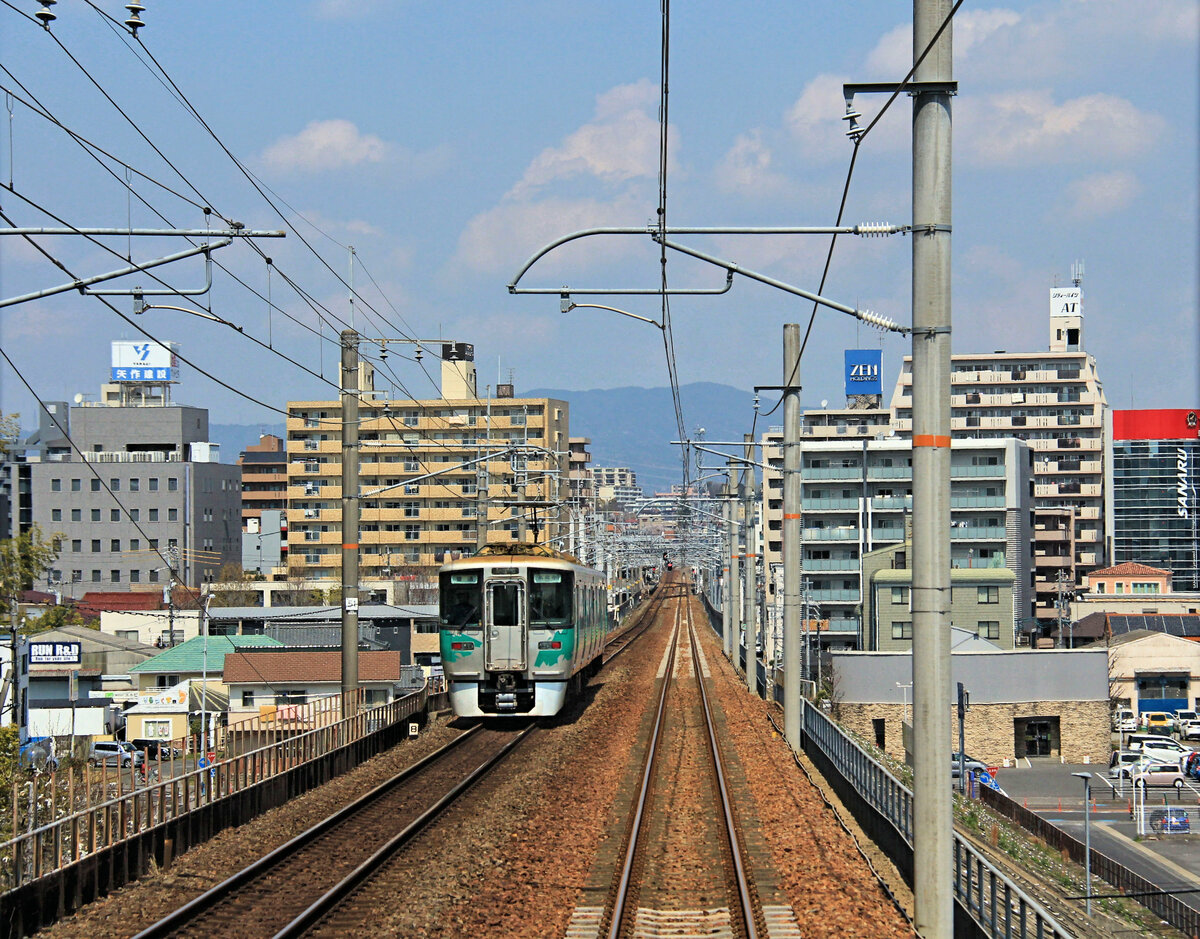 Die Aichi Ringbahn bedient im südlichen Teil vor allem die vielen Siedlungen um die Toyota Autofabrik herum. Im nördlichen Teil haben sich zahlreiche Schulen und Universitäten angesiedelt, ebenso hat sich dieses Gebiet mit vielen hübschen kleinen Häuschen zu einem Wohnort für Personen aus der städtischen Agglomeration Nagoya entwickelt. So gehört die Aichi Ringbahn zu den wenigen profitablen Bahnen in Japan. Ihre ursprüngliche Aufgabe, den Güterverkehr zwischen den von Nagoya ausstrahlenden Linien um die Stadt herumzuführen, hat sie nie wahrgenommen. Im Bild Zug 2110 im dicht besiedelten Gebiet der Stadt Toyota. 4.April 2020 