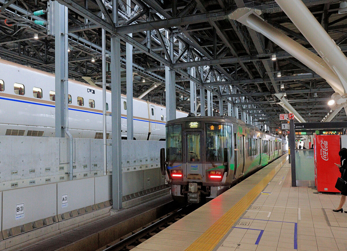 Die Ainokaze Toyama Bahn, eine der drei neu entstandenen Privatbahnen im Hokuriku-Gebiet: Hokuriku («Nord-Land») bezeichnet die im Winter extrem schneereiche Küste dem Japanischen Meer entlang (von der alten Kaiserstadt Kyoto aus gesehen) in den Norden; gemeint sind meist die 3 Praefekturen Fukui, Ishikawa (mit der Hauptstadt Kanazawa), und Toyama. Der Hochgeschwindigkeitszug Shinkansen erreichte die Stadt Kanazawa via Toyama von Tokyo aus am 14. März 2015. Mit der Eröffnung dieses Hokuriku Shinkansen wurde die alte JR Linie der Küste entlang innerhalb der Praefektur Toyama zur privaten Ainokaze Toyama Bahn. Diese Bahn behielt ihre bisherige JR-Infrakstruktur und erhielt von der JR 16 Züge Serie 521; dazu beschaffte sie bis heute 6 weitere neue Züge Serie 521. Im Bild: Im neuen Bahnhof Toyama wartet der Ainokaze Toyama Bahn Zweiwagenzug 521 Nr. 18 auf Abfahrt nach Kanazawa. Darüber ist ein Shinkansen Hochgeschwindigkeitszug aus Tokyo zu sehen. 19.März 2024 