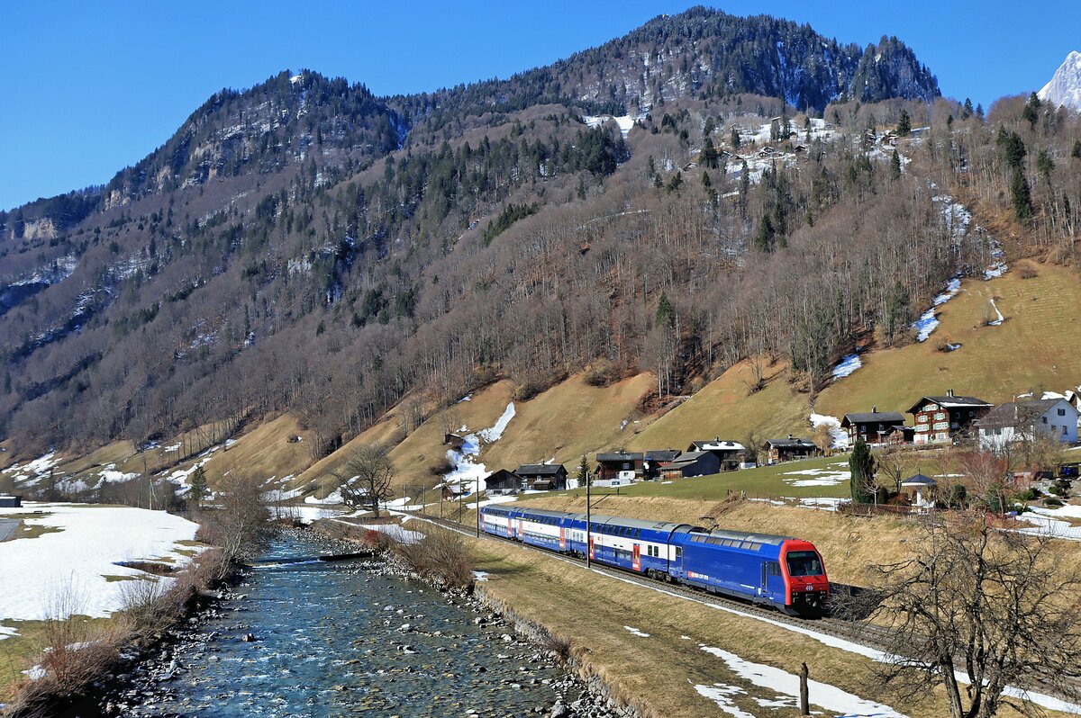 Die Bahn im Kanton Glarus: Luchsingen ist eines der wenigen Dörfer, wo noch eine Siedlung mit typischen alten Holzgebäuden steht. Linth-aufwärts fährt daran vorbei ein S-Bahnzug aus Zürich, gestossen von Re 450 084. 28.Februar 2022 