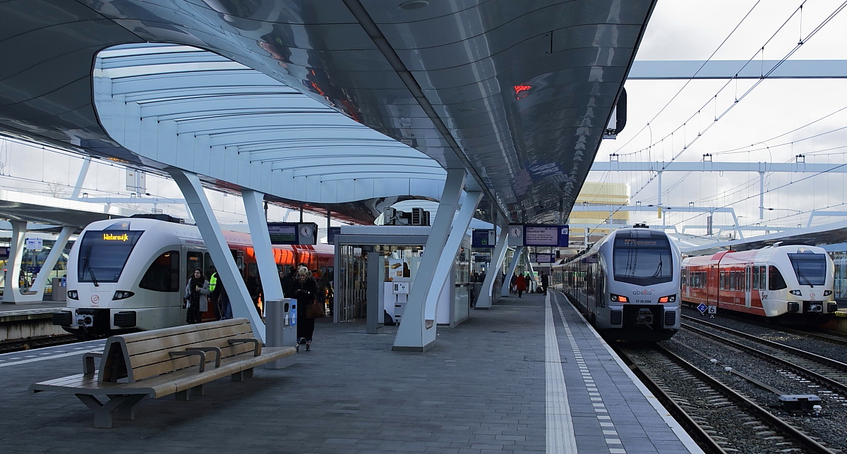 Die Bahnsteigüberdachungen von Arnhem Centraal nehmen die geschwungenen Formen des Bahnhofsgebäudes auf. Durch die verglasten Öffnungen fällt Tageslicht auf die Bahnsteige. Am 02.01.2018 steht der Abellio-ET 25 2304 (RE 19 Arnhem Centraal - Düsseldorf Hbf.) zwischen zwei Veolia-GTW 2/6. 