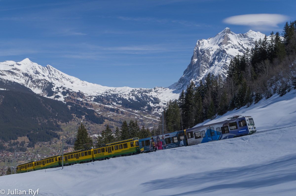 Die BDhe 4/8 132 und 134 fahren mit einem (leider nicht sehr kreativ) mit Werbung versehenen Gelenksteuerwagen am 28.03.2015 zwischen Alpiglen und Brandegg talwärts. Im Hintergrund das Wetterhorn und die Grosse Scheidegg. 
