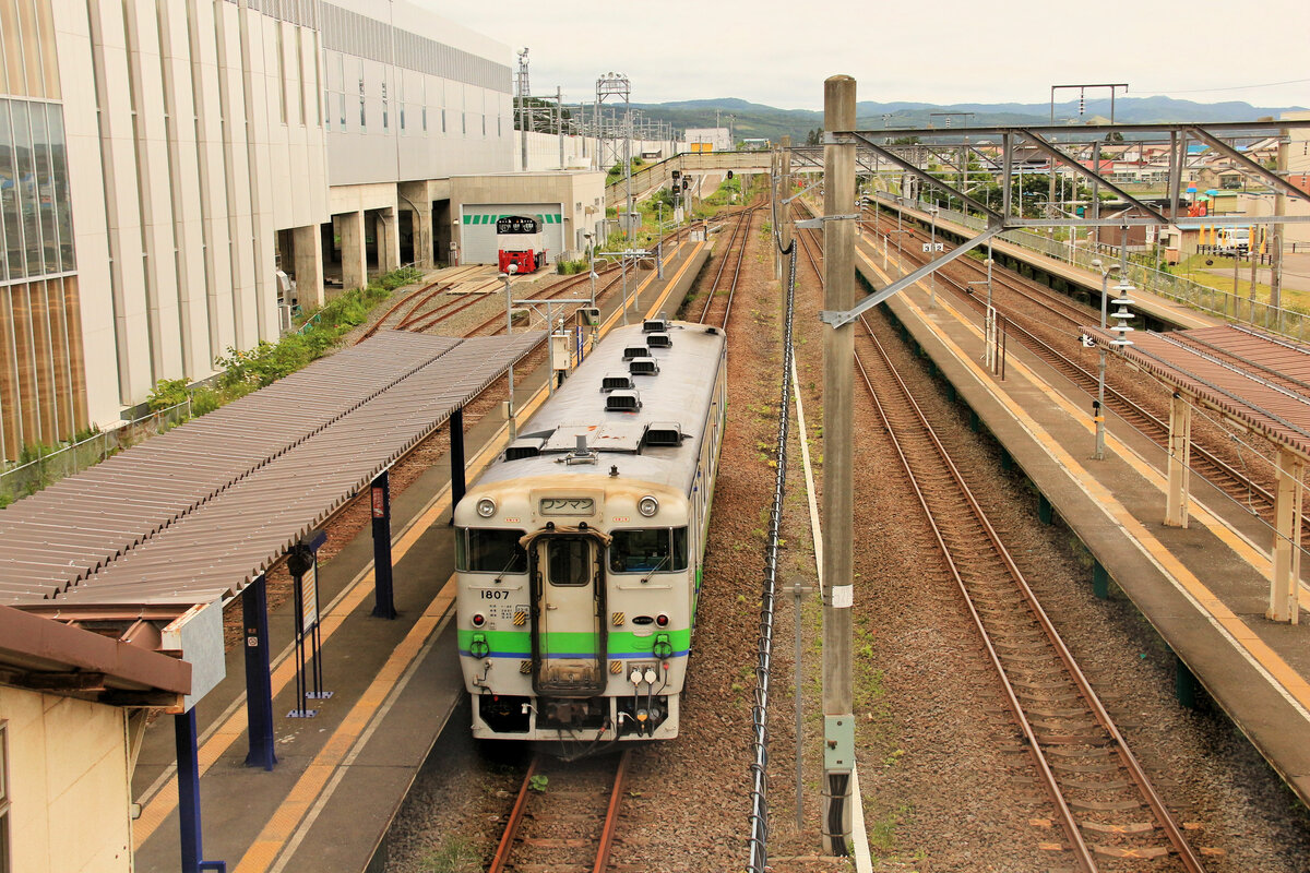 Die Dônan Isaribi-Bahn: Ursprünglich kamen die Fahrgäste zur Nordinsel Hokkaidô von der japanischen Hauptinsel Honshû her nach 4-stündiger Fahrt mit der Fähre in der Stadt Hakodate an. Von Hakodate in den Südwestzipfel von Hokkaidô führten Lokalstrecken. Als dann der Unterseetunnel (der Haupttunnel ist 53,85 km lang) am 13.März 1988 eröffnet wurde, erreichte die Unterseelinie von Honshû her als erstes die kleine Ortschaft Kikonai an einer dieser Lokalstrecken im Südwesten. Für die Intercity- und schweren Containerzüge, die jetzt nach Hokkaidô kamen, wurde die Strecke von Kikonai zum weiter östlich gelegenen Hakodate entsprechend elektrifiziert. Als dann die Shinkansen-Hochgeschwindigkeitsstrecke nach Hokkaidô eröffnet wurde (26.März 2016), versah man den Unterseetunnel mit einem dritten Gleis, damit sowohl der Shinkansen (1435 mm Spur) wie auch die Containerzüge (1067 mm Spur) durchfahren können. Gleichzeitig wurde die ehemalige Hauptstrecke Kikonai-Hakodate, die nun für den Personenverkehr durch den Shinkansen (nach Tunnelausgang auf einer ganz neuen Strecke) ersetzt wurde, an eine v.a. lokal geprägte Aktiengesellschaft - eben die Dônan Isaribi-Bahn - abgegeben, die die wenigen Dörfer zwischen Kikonai und Hakodate bedient. Diese Strecke wird aber jetzt hauptsächlich für die Containerzüge verwendet. Im Bild sieht man die riesigen neuen Bahnhofsgebäude für den Shinkansen, und darunter recht verlassen einen Wagen der Dônan Isaribi Bahn; es handelt sich um den ehemaligen JR-Wagen KIHA 40 1807, noch im alten Anstrich von JR Hokkaidô. Kikonai, 24.Juli 2016  