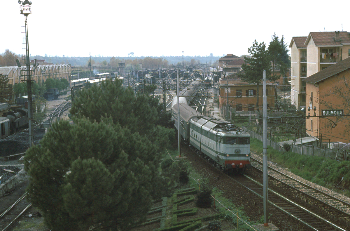 Die E646 105 fährt mit dem am frühen Abend verkehrenden Rapido Pescara - Roma aus dem Bahnhof Sulmona aus. Der Zug besteht ausschließlich aus Waggons des Typs UIC-X, die sich damals noch in der Ablieferung befanden. Das Gleis rechts neben der Lokomotive gehört zur Eisenbahn Sulmona - Carpinone - Isernia, die heute nur noch von Sonderzügen der Fondazione FS befahren wird. Aber damals stand noch ein Dieseltriebwagen für diese Strecke bereit, im Hintergrund rechts neben dem Zug. Links ist das Deposito Locomotive (Bahnbetriebswerk), ganz am Bildrand stehen eine Dampflok und ein Heizwagen vor einem Kohlevorrat. April 1980, Canon AE1, Canoscan, GIMP