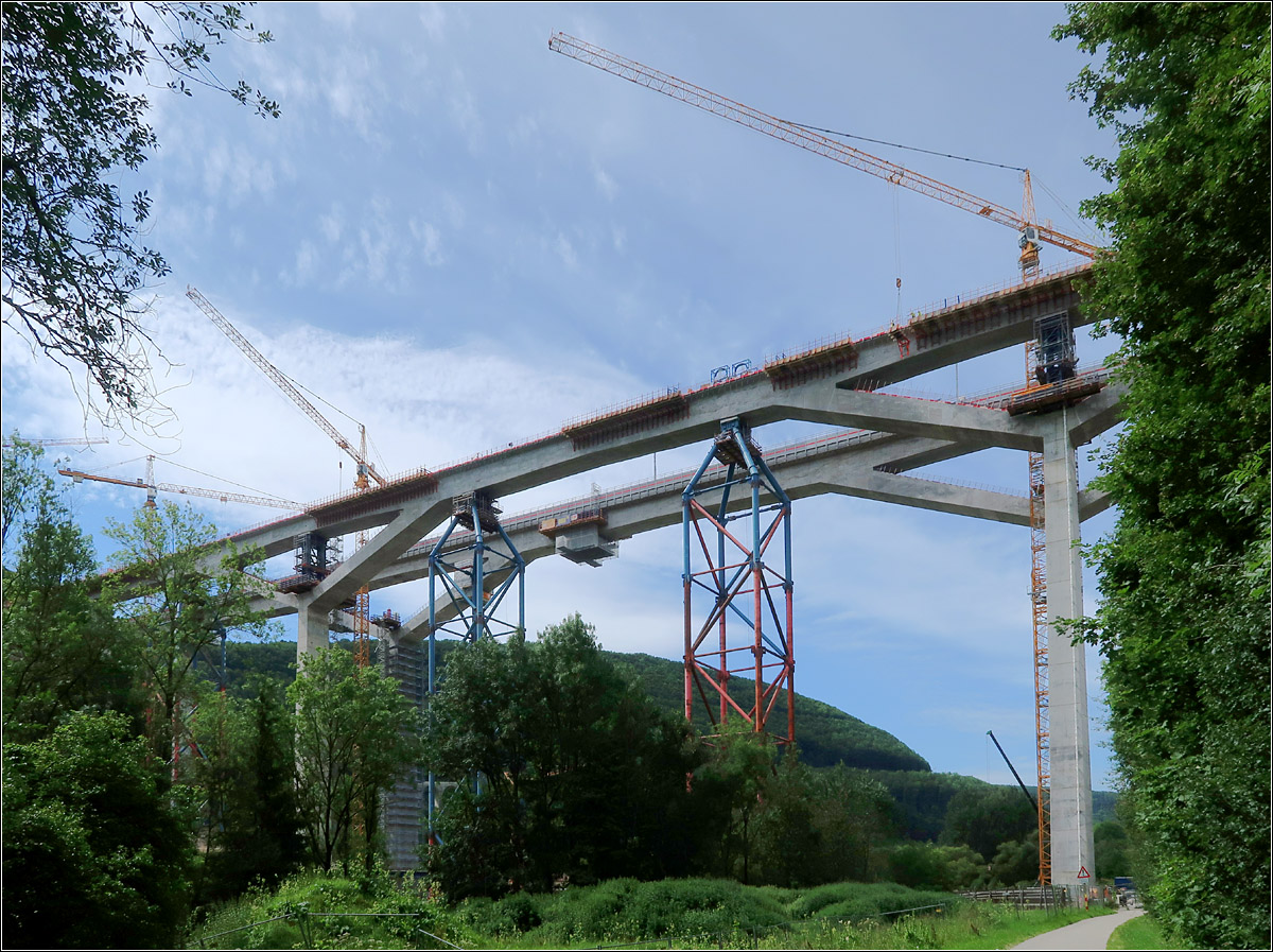 Die Filstalbahnbrücke -

... der Neubaustrecke Stuttgart - Ulm. Noch stehen an der vorderen Brücke Hilfstützen aus Stahl aber schon ab 11. Dezember 2022 sollen hier Züge fahren. Kürzlich wurde der Brückenschlag auch der zweiten Brücke gefeiert.

Die beiden Brücken sind 85 m hoch und 485 m bzw. 472 m lang. Sie liegen im Bereich des Albaufstiegs zwischen dem Boßlertunnel (8,8 km) und dem Steinbühltunnel (4,8 km). Die Steigung beträgt ca. 2,5 %.

Die Brücken wurde möglichst filigran mit den flachen Y-Pfeilern gestaltet. Ungewöhnlich ist die monolithische Bauweise, die Stützen und der Oberbau sind fest miteinander verbunden, ohne Lager.

Der Weg unten rechts befindet sich auf der Trasse der früheren Tälesbahn Geislingen/Steige - Wiesensteig.

10.09.2021 (M)