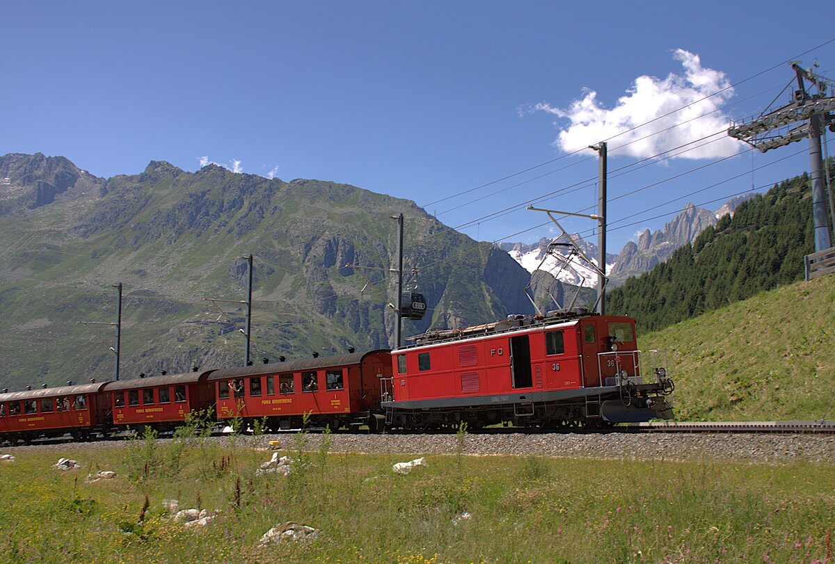 Die FO 36, �ber 80 Jahre alt, zieht am 04.07.2026 den Jubil�umszug, im Rahmen der Feierlichkeiten zu 100 Jahre Furka-Oberalp-Bahn, von Andermatt her zum Oberalppass. Aufgenommen habe ich das Bild kurz vor N�tschen. Im Hintergrund ist die Bergwelt ums G�schneralptal zu sehen.
Die im Zug verwendeten Wagen geh�ren der DFB.
Jahrzehntelang zogen diese Lokomotiven den langsamsten Schnellzug der Welt, den Glacier-Express. In der langen Zeit wurde daf�r verschiedenstes Wagenmaterial verwendet. Bis zur Einf�hrung der Triebwagen bef�rderten diese Maschinen auch die Personenz�ge.