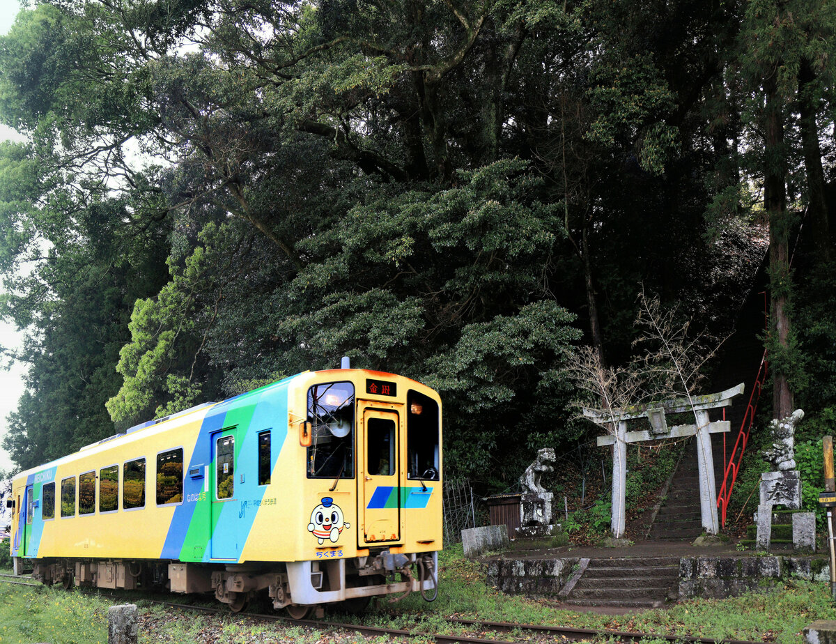 Die Heisei Chikuhô Bahn im Nordosten der Insel Kyûshû: Unmittelbar vor der Aufstiegstreppe zum Heiligtum in der Höhe oben fährt Triebwagen 406 vorbei. Das Eingangstor wird links und rechts von zwei tanzenden Löwen bewacht. 16.April 2023 