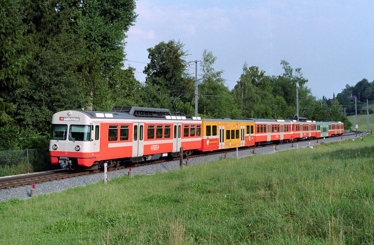 Die RBS-Be 4/12 60 und 53 am 22. Juli 2004 auf der damaligen Linie W bei Bolligen. Der Be 4/12 60 trägt einen Versuchsanstrich mit einer auf Seite Worb weiss gestalteten Front. Auf dem gelben Niederflur-Mittelwagen B 42 wurden im Rahmen einer künstlerischen Gestaltung von Judith Zaugg Comic-Figuren angebracht.