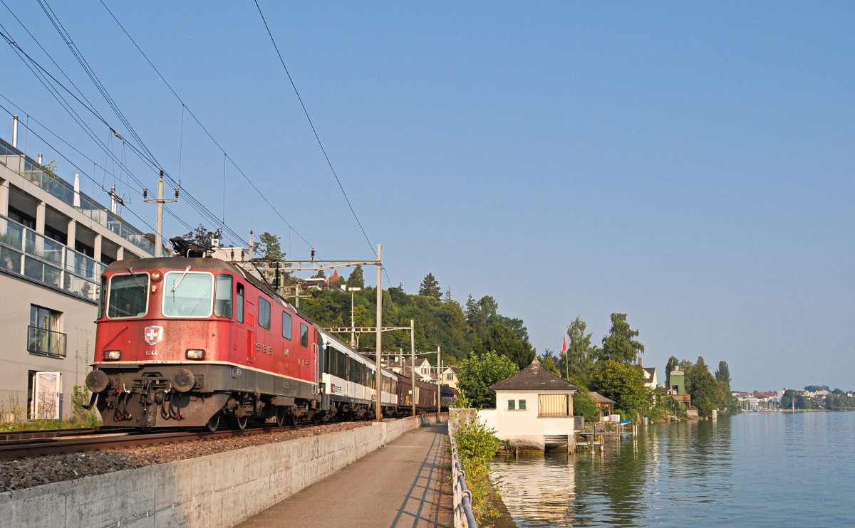 Die Re 4/4 II 11244 ist mit zwei Gepäckwagen (ex SNCF) sowie drei gedeckten Güterwagen bei Wädenswil dem Zürichsee entlang unterwegs. Aufgenommen am 12.08.2015