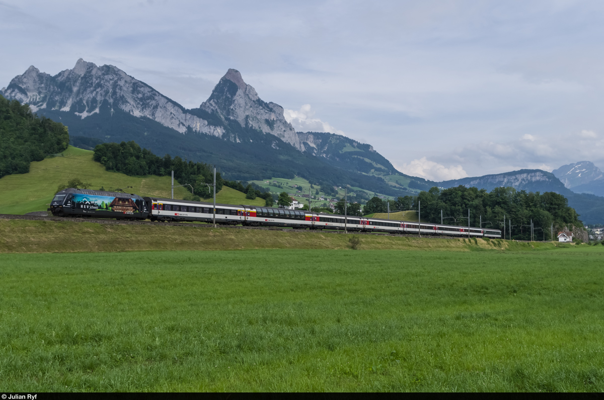 Die Re 460 023 mit Werbung für den Hauseigentümerverband Schweiz HEV  bespannte am 29. Juni 2015 einen IR am Gotthard. Hier fährt sie kurz vor Steinen nordwärts. Im Hintergrund die Mythen.