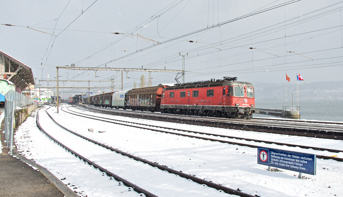 Die Re 620 064-6 ist mit einem Ferngüterzug RBL-Buchs SG bei Schneefall und Sonnenschein entlang des Zürichsees unterwegs. Wädenswil, 07.04.2021.