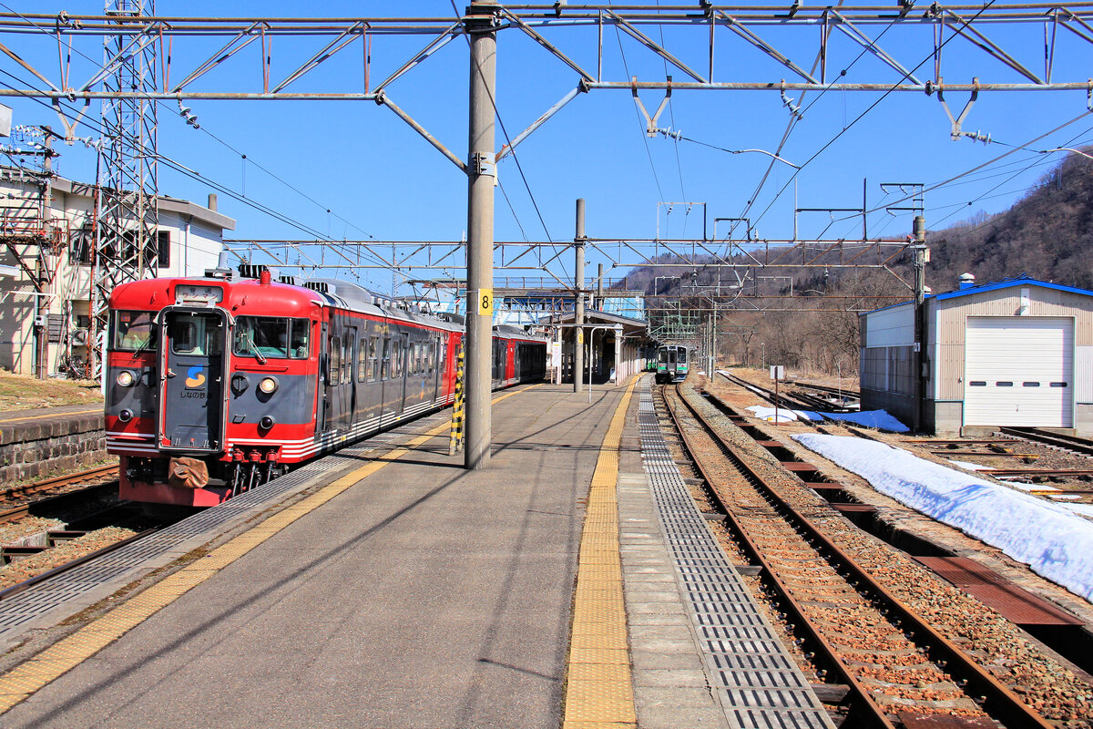 Die Shinano-Bahn (II): Als die Hochgeschwindigkeitsbahn (Shinkansen) von Nagano an die K�ste des Japanischen Meeres nach Toyama und Kanazawa verl�ngert wurde (Er�ffnung 14.M�rz 2015), wurde die Passstrecke �ber die My�k�-Hochebene bis auf einen geringen Lokalverkehr �berfl�ssig. G�terverkehr gab es hier schon lange nicht mehr. Diese Passstrecke bildete einst die zweite Ueberquerung der Alpen auf der Reise von Tokyo ans Japanische Meer zu den St�dten Toyama und Kanazawa (die erste Alpen�berquerung war die Usui-Steilrampe und dann der Abstieg von Karuizawa nach Nagano). Die nun auch �berfl�ssig gewordene zweite Passstrecke wurde dann so aufgeteilt, dass die Shinano-Bahn (hier als Nord-Shinano Bahn bezeichnet) die Strecke bis zur Praefekturgrenze auf dem My�k� Pass �bernahm, und die von der anschliessenden Praefektur Niigata getragene Echigo Tokimeki Privatbahn die Fortsetzung ans Japanische Meer hinunter. So muss man jetzt auf der My�k� Hochebene (My�k� K�gen) umsteigen. Die Nord Shinano-Bahn ist 37, 3 km lang. - Im Bild die  Grenzstation  My�k� K�gen ( My�k� Hochebene ), im Vordergrund f�hrt Shinano Bahn Triebzug S1 zur�ck in die Praefektur Nagano, w�hrend rechts ein Zug der Echigo Tokimeki Bahn Passagiere aufgenommen hat, die die Reststrecke ans Meer hinunter fahren wollen. 9.M�rz 2020  