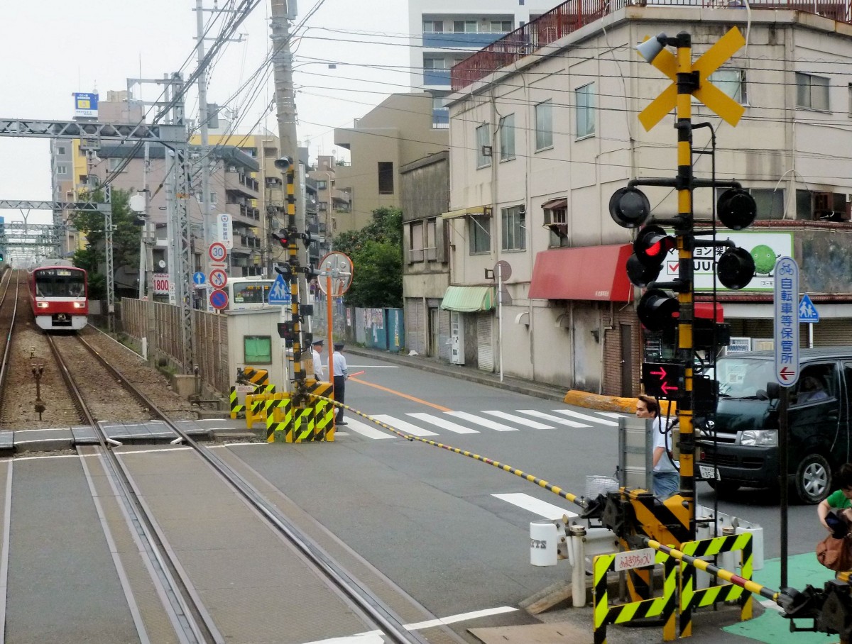 Die Stammstrecke des Keihin Kyûkô (Keikyû)-Konzerns: Am selben Ort wie das Bild mit dem blauen Zug 2157 in Hatchô Nawate, Stadt Kawasaki. Blick auf die Bahnschrankenanlage; bei allen Bahnschranken in Japan wird mit Pfeilen die Richtung angegeben, aus der sich der Zug nähert - hier im Bild leuchten beide Pfeile. Der Gegenzug ist der Achtwagenzug 1613. 25.Juni 2011.  