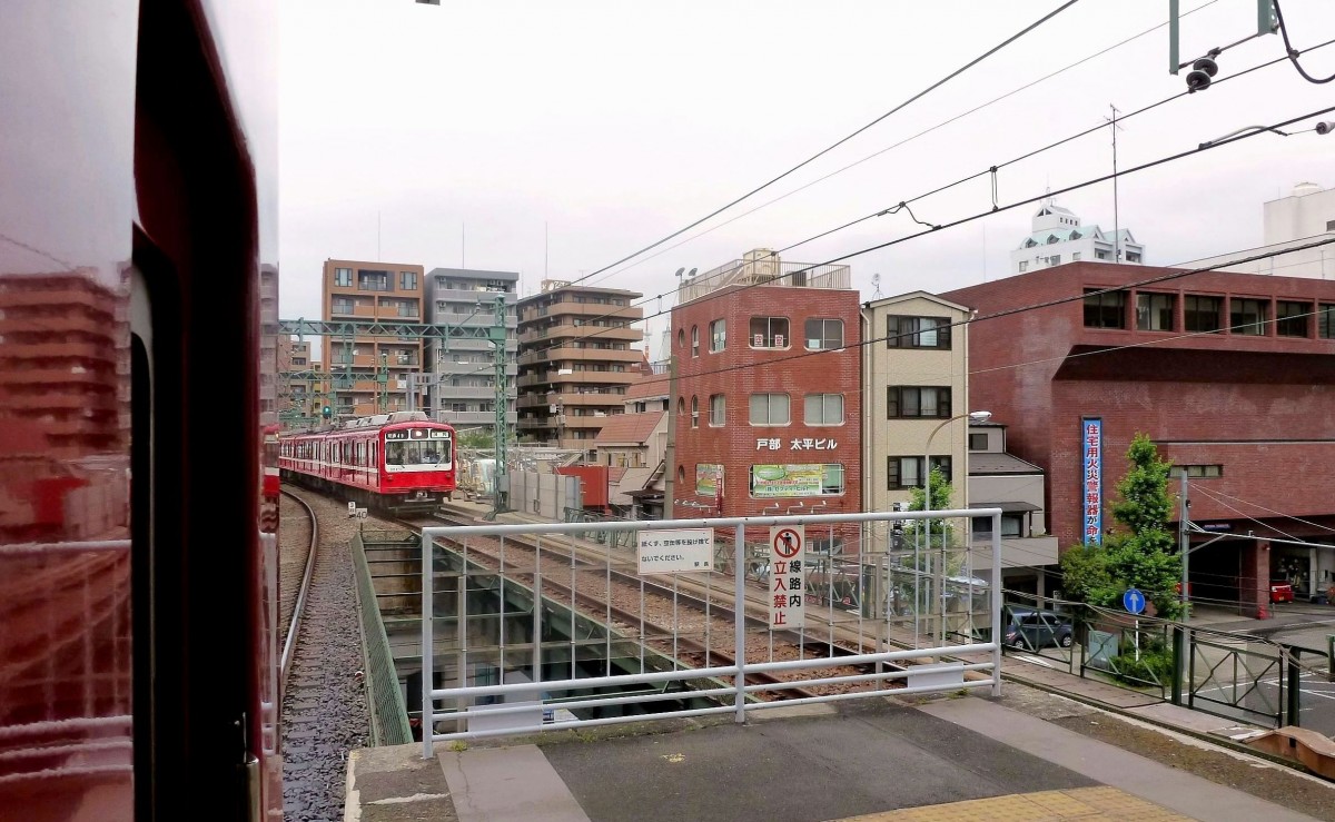 Die Stammstrecke des Keihin Kyûkô (Keikyû)-Konzerns: Häuserblöcke so weit das Auge reicht. Warnschilder an der Bahnsteigabschrankung weisen auf die Gefahren hin, den Bahnkörper zu betreten und bitten, hier keinen Müll und keine leeren Dosen hinabzuwerfen. Bild aus einem wartenden Zug; Zug 821 (Serie 800) fährt in die Station Tobe in Yokohama ein, 25.Juni 2011.   