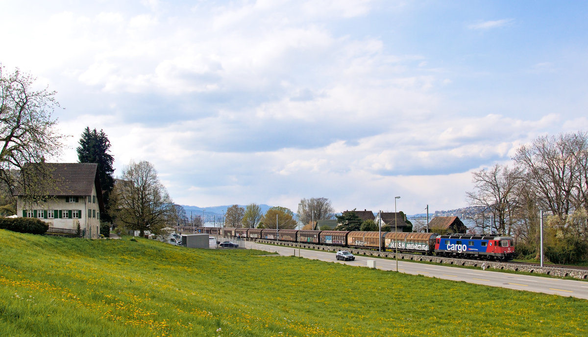 Die Ufer des Zürichsees, welcher im Hintergrund zu sehen ist, sind fast lückenlos mit Gewerbe- und Wohnsiedlungen überbaut. In einer dieser Lücken ist die Re 620 023  Rupperswil  mit einem Güterzug unterwegs. Aufgenommen am 15. April 2021 bei Au ZH.
