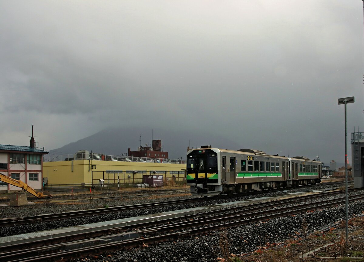 Die ursprüngliche Hauptlinie vom Fährhafen Hakodate zur Hauptstadt von Hokkaidô, Sapporo, führte über den Pass von Kutchan; jetzt wird diese direkte Linie nur noch von Lokalzügen befahren. Der Höhepunkt dieser Strecke ist der mächtige, ca. 1900 Meter hohe Vulkan Yôteizan. Es seien vier Versuche gezeigt, den Yôteizan irgendwie zu erwischen. In einem nahenden Regensturm fahren die beiden Triebwagen H100-7 und H100-4 von Kutchan aus Richtung Otaru. 29.Oktober 2022   