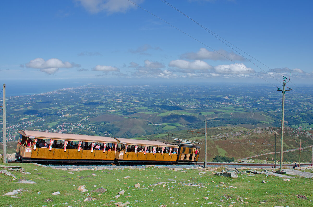 Dienstag den 19.09.2023 um 14:09 Uhr in Ascain (F). Aussicht bei der Bergstation La Rhune. Die Petit Train de la Rhune fährt die Gäste von der Talstation Col de Saint-Ignace zur Bergstation. Hier kommt soeben einer dieser Züge Bergwärts zur Bergstation. Angaben zur Bahn; Streckenlänge: 4,2 km, Spurweite: 1'000 mm, Stromsystem: 3'000 V 50 HZ, Maximale Neigung: 250 Promille, Zahnstangensystem: Strub, Höchstgeschwindigkeit: 9 km/h, Höhe Talstation: 170 müM, Höhe Bergstation: 888 müM, Lokomotiven: SLM. Im Hintergrund befindet sich der Golf von Biskaya (Atlantische Ozean). Koordinaten GMS (Grad, Minuten, Sekunden): N 43° 18’ 32.1’’ O 1° 38’ 2.9’’