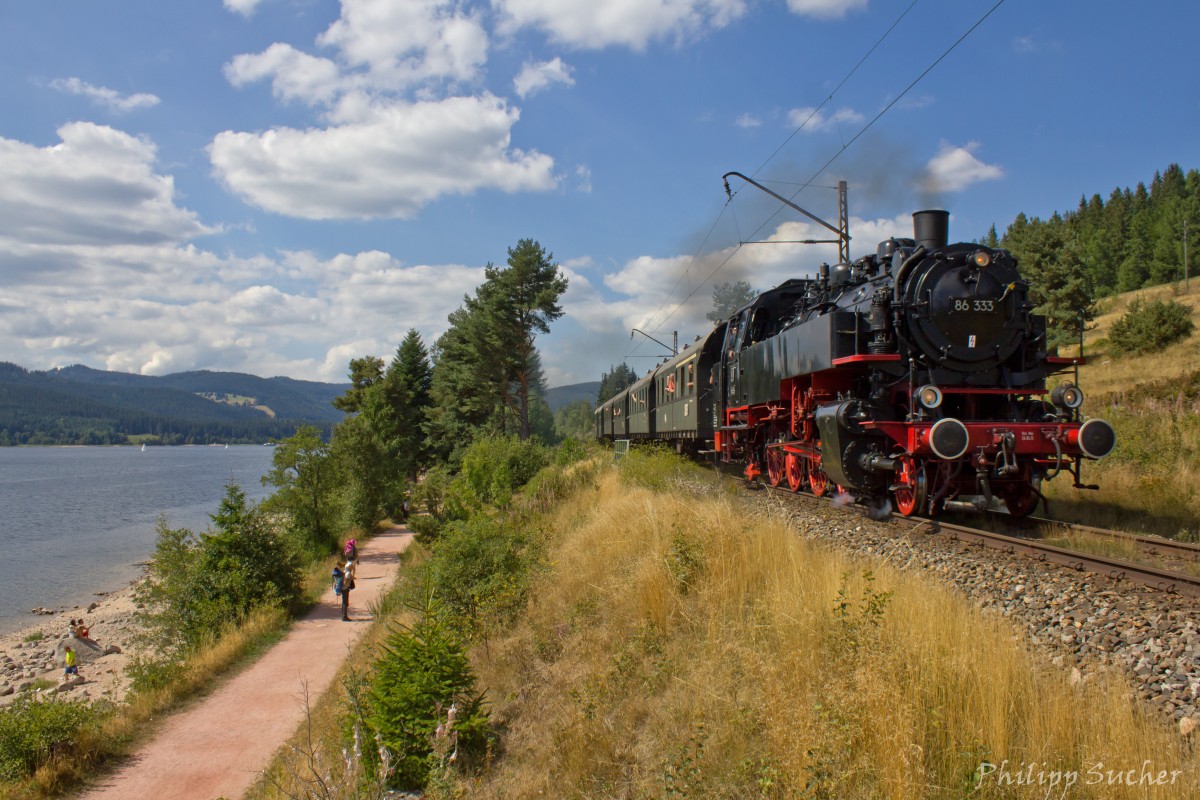 Direkt am Schluchsee entlang verläuft die Dreiseenbahn bei Aha. 86 333 ist hier am 22.08.2015 mit dem Museumszug der IG 3-Seenbahn unterwegs nach Seebrugg, während die zahlreichen Wanderer andächtig stehen bleiben und der Lok hinterherblicken.