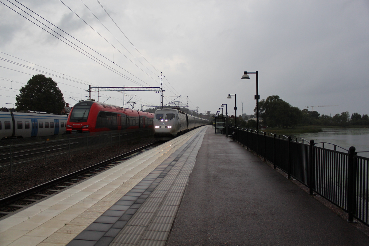 Direkt am See Frösjön liegt der Bahnhof Gnesta. Am Nachmittag des 10.09.2018 zog ein heftiges Gewitter über die Region hinweg. Links steht ein X60 als Pendeltåg nach Södertälje bereit, während in der Mitte ein X74 als mtr Express nach Göteborg durchfährt. Rechts ist ein X2 als Snabbtåg 536 nach Stockholm unterwegs.