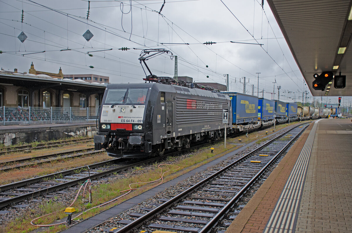 Donnerstag den 20.04.2023 um 10:08 Uhr in Basel Bad Bf. Ein Intermodal (Intermodaler Verkehr umfasst den Transport von Gütern in ein und derselben Ladeeinheit oder demselben Strassenfahrzeug mit zwei oder mehreren Verkehrsträgern, wobei ein Wechsel der Ladeeinheit, aber kein Umschlag der transportierten Güter selbst erfolgt) wird von der 91 80 6189 991-3 D-DISPO Class 189-VE durch den Badischen Personenbahnhof gezogen. Bezeichnung: SBB Re 474, bzw. ES 64 F4-091. Hersteller: SIEMENS. Baujahr: 2004. Fabriknummer: 20739. Typ: ES64F4. Leistung: 6'400 kW. Wetter: Trüb und Regen.