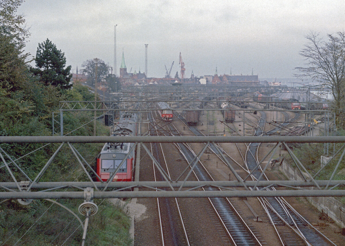DSB Bahnhof Helsingør: Im Oktober 1985 waren die Vorbereitungen für den elektrischen Betrieb der Kystbanen in vollem Gang. Eines Nachmittags im Oktober machte ich diese Aufnahme, die Aspekte des damaligen regen Bahnlebens in Helsingør zeigt: Etwas undeutlich ist die Elektrolokomotive EA 3004 (Scandia/BBC 1985) links zu sehen, weiter weg befindet sich ein DSB-Dieseltriebzug (ML + FL + ... + ML), viele Güterwagen warten auf die Weiterfahrt, rechts im Bild steht eine dieselelektrische Lokomotive des Typs ME mit einem Güterzug, und im Fährhafen liegt eine Eisenbahnfähre. - Scan eines Farbnegativs. Film: Kodak CL 200 5093. Kamera: Minolta XG-1.