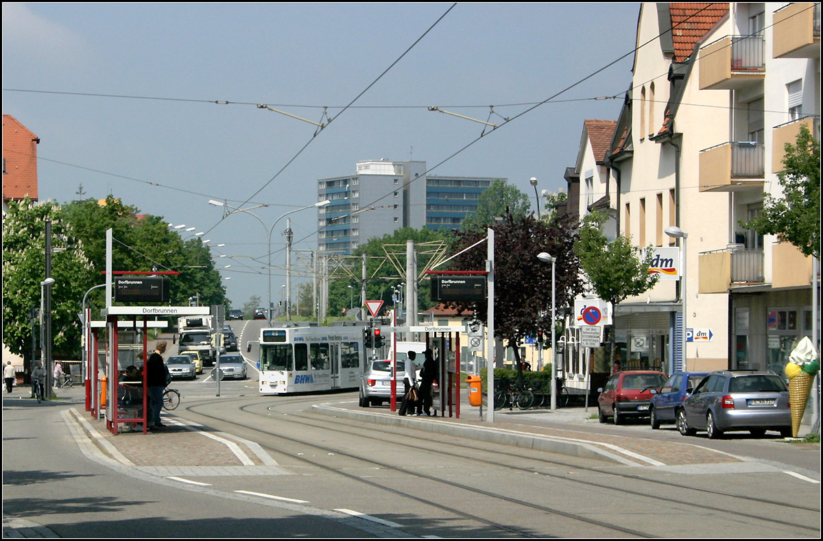 Durch Freiburg-Haslach ins Rieselfeld -

Die Haltestelle 'Dorfbrunnen' in Haslach. Die GT8Z-Tram kommt nicht aus einer Seitenstraße sondern schlängelt sich aus dem eigenen Bahnkörper entlang der Opfinger Straße in die Straßenmitte Carl-Kistner-Straße. In der Ortsdurchfahrt von Haslach wurde eine Einfachfahrleitung verwendet, die erfreulicherweise meist an den Gebäuden aufgehängt wurde, so entfallen störende Masten.

11.05.2006 (M)