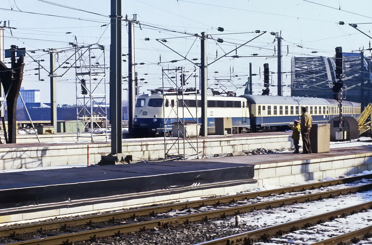 E 10 mit einer schönen Garnitur Reisezugwagen in oceanblau/beige bei der Einfahrt in den Kölner HBF. Die Aufnahme entstand ca. Januar 1990 (Scan vom Dia, ggfs. auch früher). Die Überdachungsverlängerung des Bahnsteiugvorfelds ist noch nicht abgeschlossen. Schöne zu sehen auf dem Bahnsteig ist ein kleiner Eisenbahner mit seinem Vater.